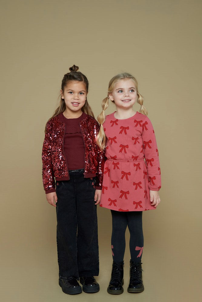 Jubel Jurk AOP - Queen Of Hearts Kind Poserend: "Kind poseert in de Jubel Jurk AOP - Queen Of Hearts met Strikjes Design in Roze, met een speelse uitstraling en comfortabele pasvorm."