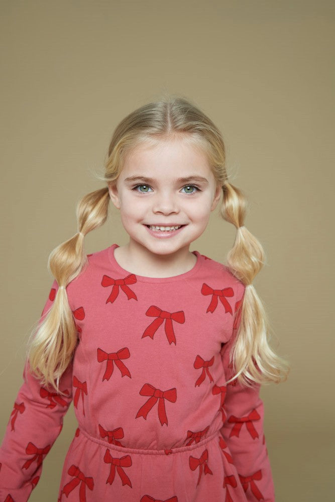 Jubel Jurk AOP - Queen Of Hearts Kind Poserend: "Kind poseert in de Jubel Jurk AOP - Queen Of Hearts met Strikjes Design in Roze, met een speelse uitstraling en comfortabele pasvorm."-