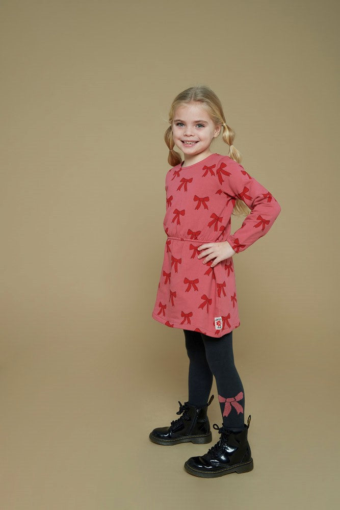 Jubel Jurk AOP - Queen Of Hearts Kind Poserend: "Kind poseert in de Jubel Jurk AOP - Queen Of Hearts met Strikjes Design in Roze, met een speelse uitstraling en comfortabele pasvorm."