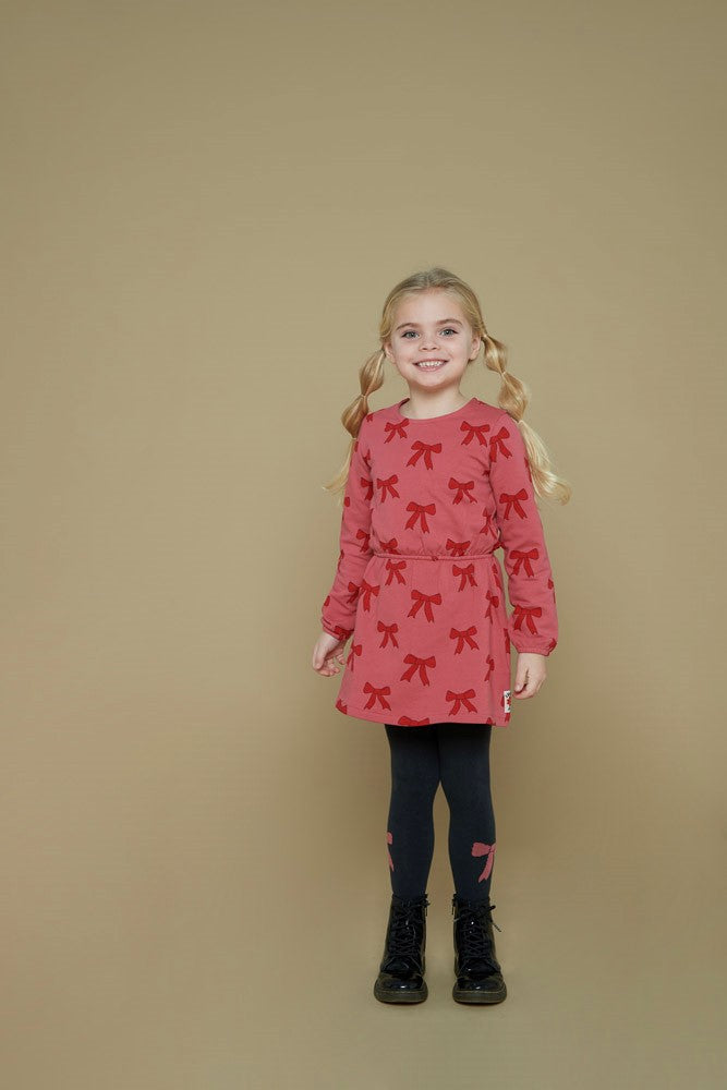 Jubel Jurk AOP - Queen Of Hearts Kind Poserend: "Kind poseert in de Jubel Jurk AOP - Queen Of Hearts met Strikjes Design in Roze, met een speelse uitstraling en comfortabele pasvorm.