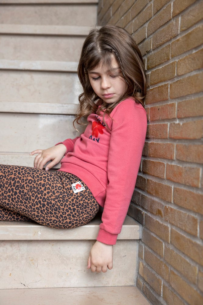 Jubel Flared Broek AOP - Queen Of Hearts Sfeerfoto: "Kind draagt de Jubel Flared Broek AOP - Queen Of Hearts in Bruin, met een speelse print van hartjes en strikjes, perfect voor casual winteroutfits."