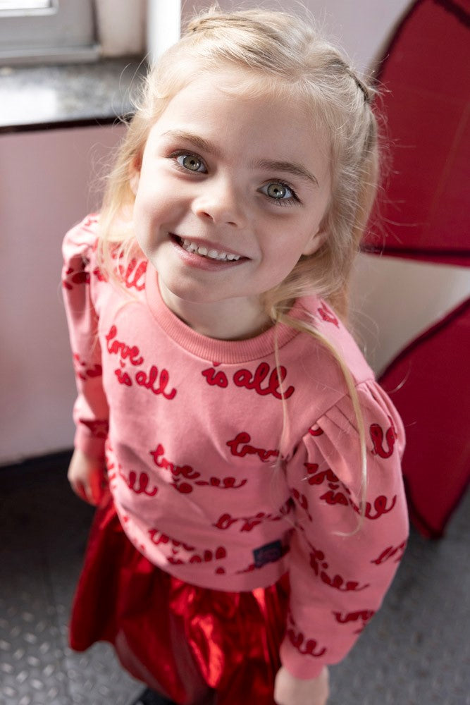 Jubel Sweater AOP - Queen Of Hearts Sfeerfoto: "Kind draagt de Jubel Sweater AOP - Queen Of Hearts in Lichtroze, met een speelse print van hartjes en strikjes, perfect voor casual winteroutfits."