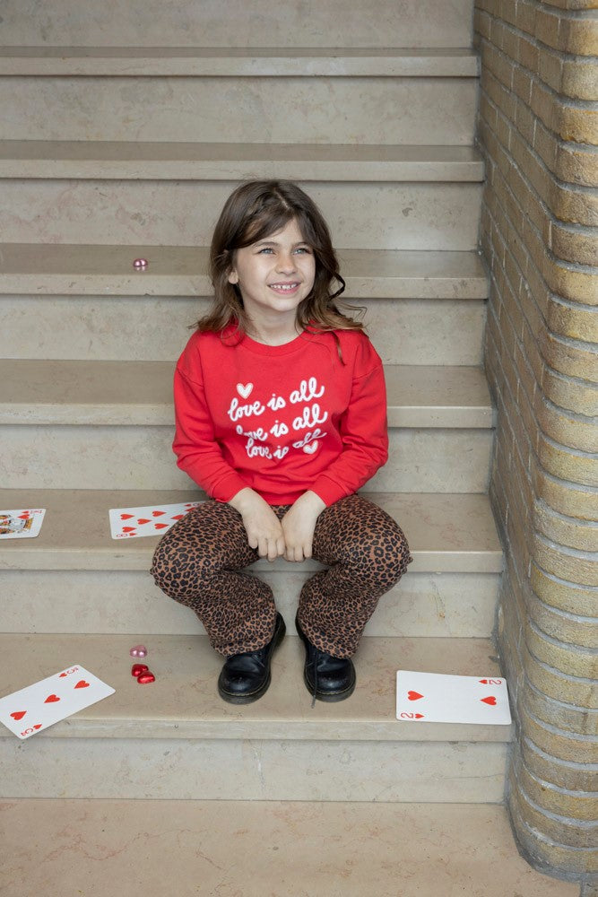 Jubel Flared Broek AOP - Queen Of Hearts Sfeerfoto: "Kind draagt de Jubel Flared Broek AOP - Queen Of Hearts in Bruin, met een speelse print van hartjes en strikjes, perfect voor casual winteroutfits."