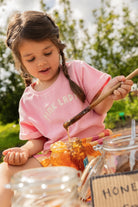 Meisje met een Sproet & Sprout Terry T-Shirt Pink Lady aan speelt met honing