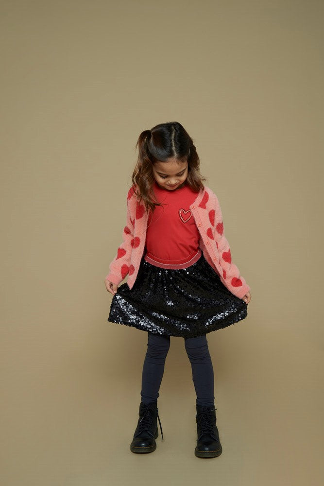 Jubel Vest Gebreid AOP - Queen Of Hearts Kind Poserend: "Kind poseert in het Jubel Vest Gebreid AOP - Queen Of Hearts in Licht Roze, met een speelse uitstraling en comfortabele pasvorm."