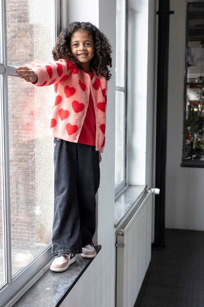 Jubel Vest Gebreid AOP - Queen Of Hearts Kind Poserend: "Kind poseert in het Jubel Vest Gebreid AOP - Queen Of Hearts in Licht Roze, met een speelse uitstraling en comfortabele pasvorm."