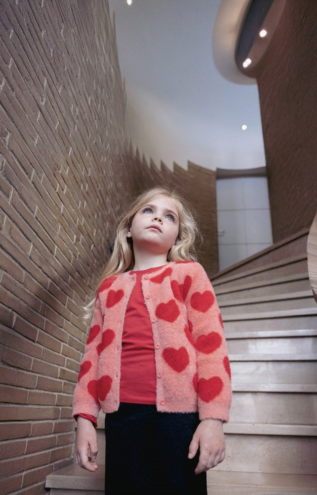 Jubel Vest Gebreid AOP - Queen Of Hearts Kind Poserend: "Kind poseert in het Jubel Vest Gebreid AOP - Queen Of Hearts in Licht Roze, met een speelse uitstraling en comfortabele pasvorm."