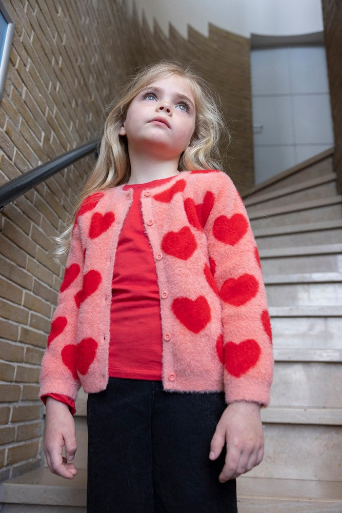 Jubel Vest Gebreid AOP - Queen Of Hearts Kind Poserend: "Kind poseert in het Jubel Vest Gebreid AOP - Queen Of Hearts in Licht Roze, met een speelse uitstraling en comfortabele pasvorm."