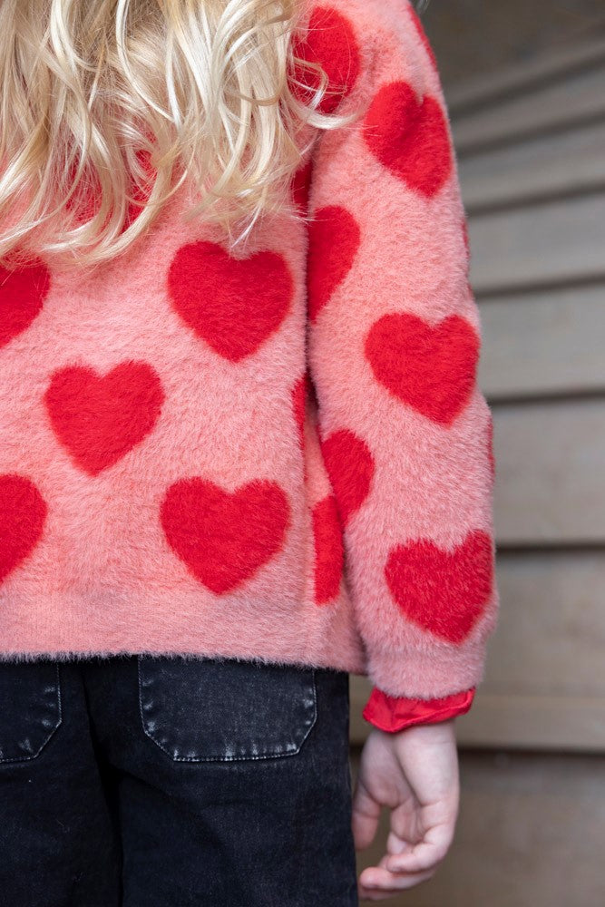 Jubel Vest Gebreid AOP - Queen Of Hearts Kind Poserend: "Kind poseert in het Jubel Vest Gebreid AOP - Queen Of Hearts in Licht Roze, met een speelse uitstraling en comfortabele pasvorm."