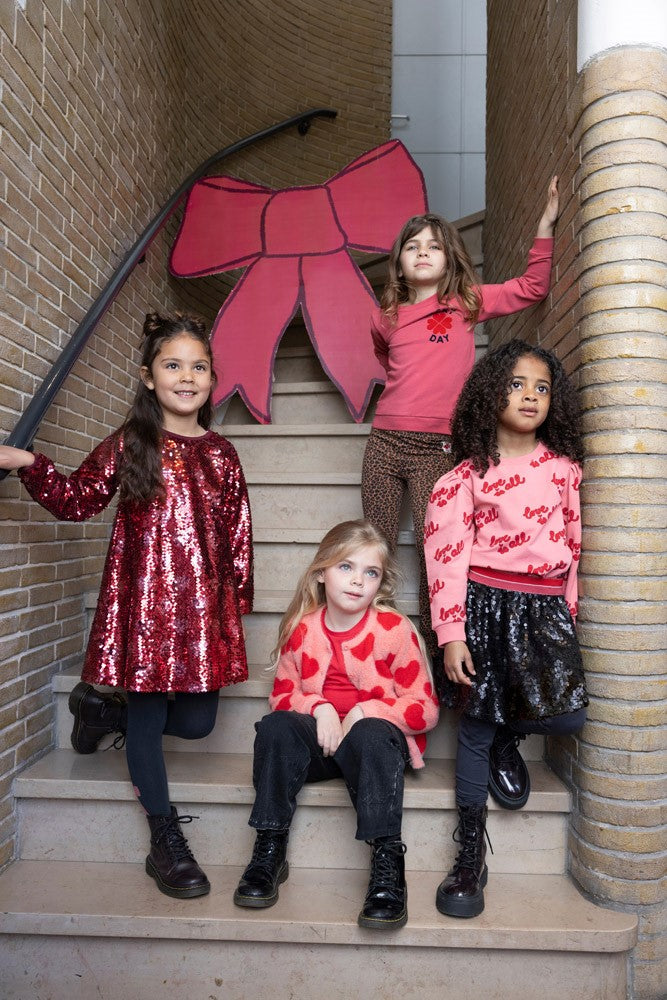 Jubel Vest Gebreid AOP - Queen Of Hearts Kind Poserend: "Kind poseert in het Jubel Vest Gebreid AOP - Queen Of Hearts in Licht Roze, met een speelse uitstraling en comfortabele pasvorm."